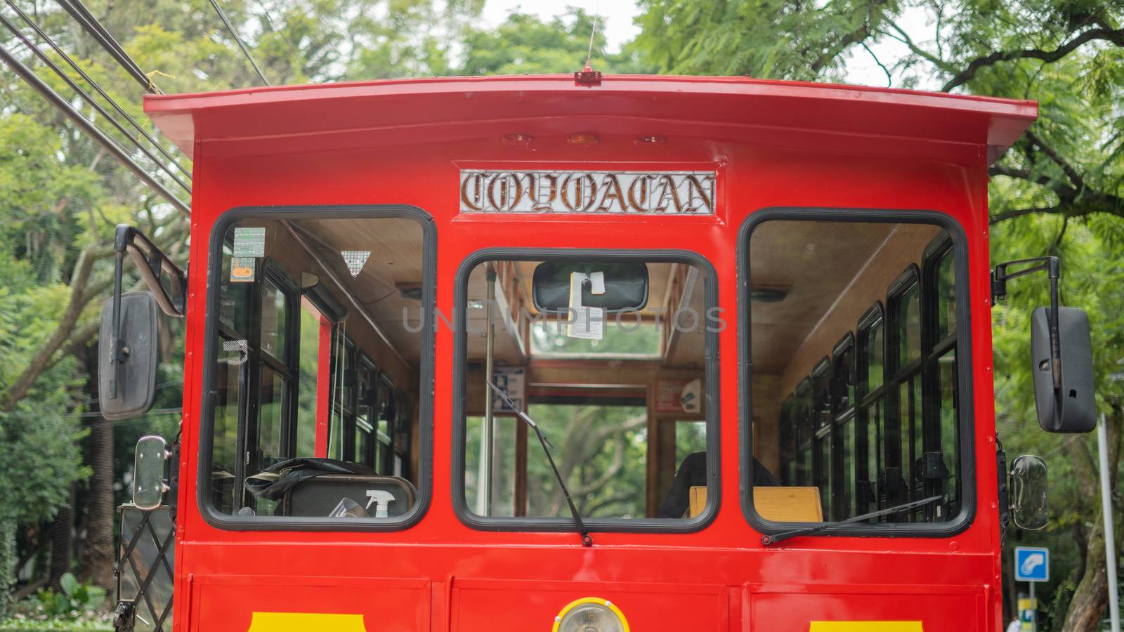 Picture of the front side of a red and yellow trolley car driving on a street surrounded by trees