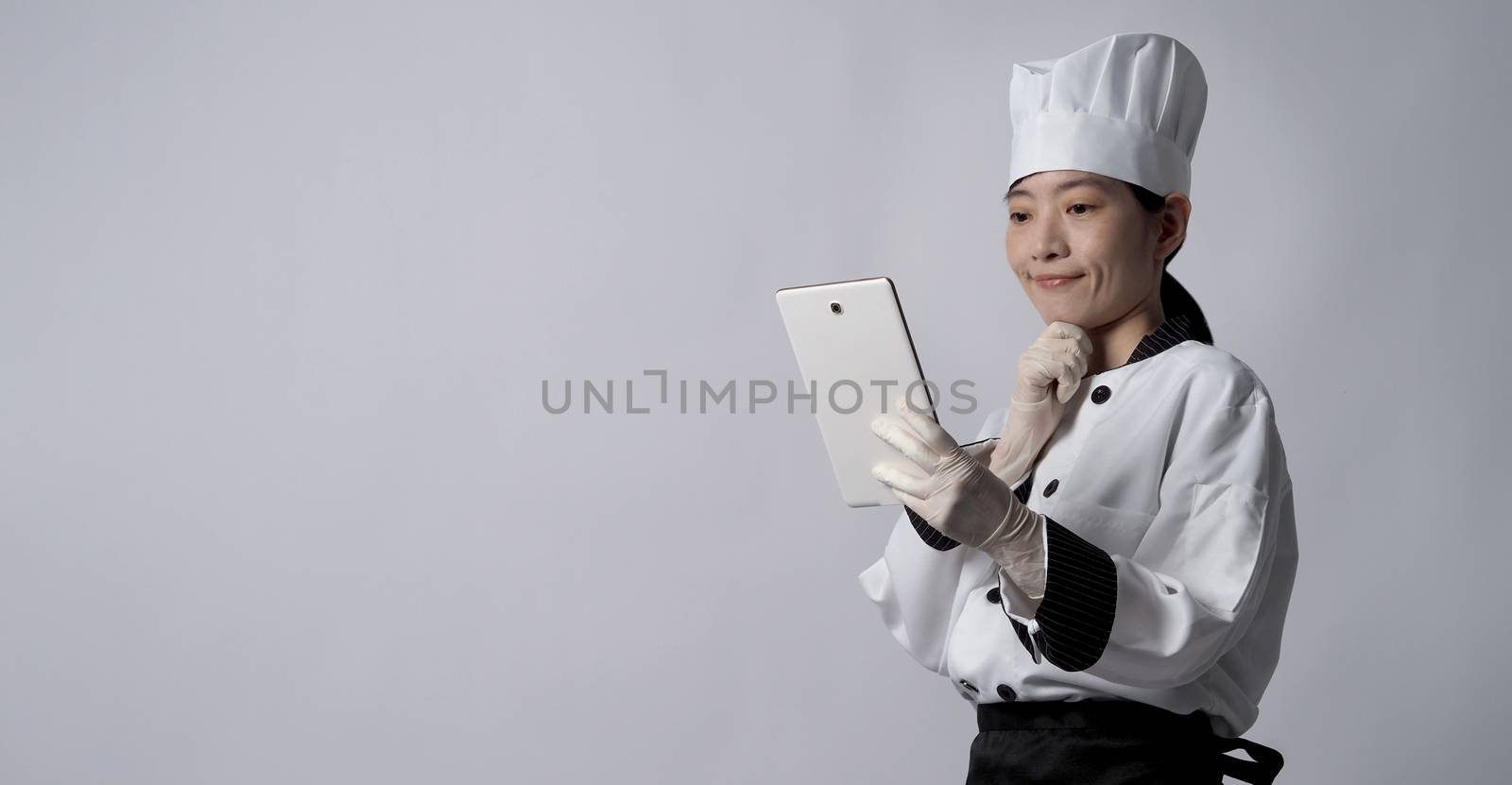 Asian woman chef holding smartphone or digital tablet and received food order from online shop or merchant application. she smiling in chef uniform and standing in studio with white color background.