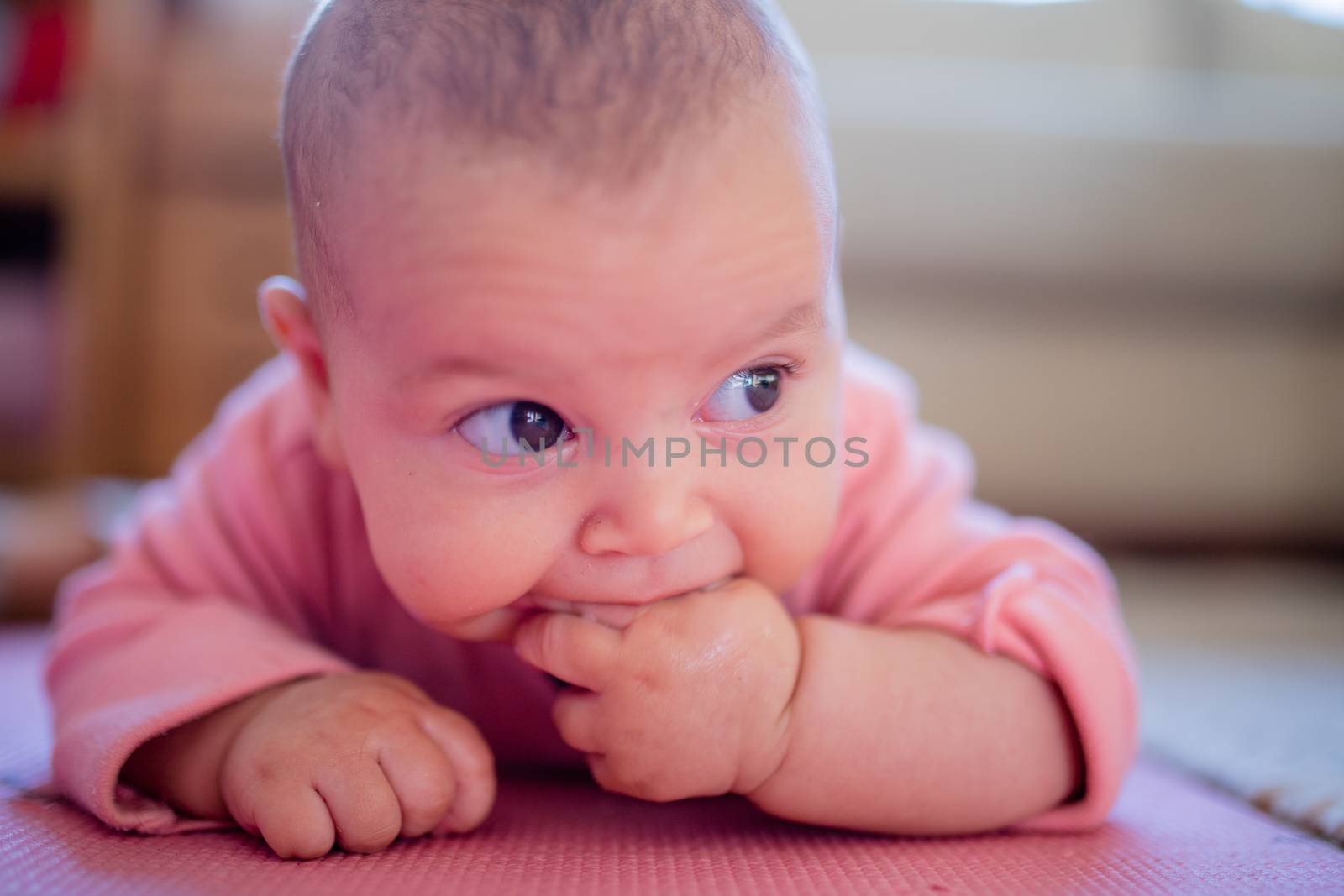 Smiling baby in a pink sleepsuit lying face down on a pink rug with two of her fingers in her mouth