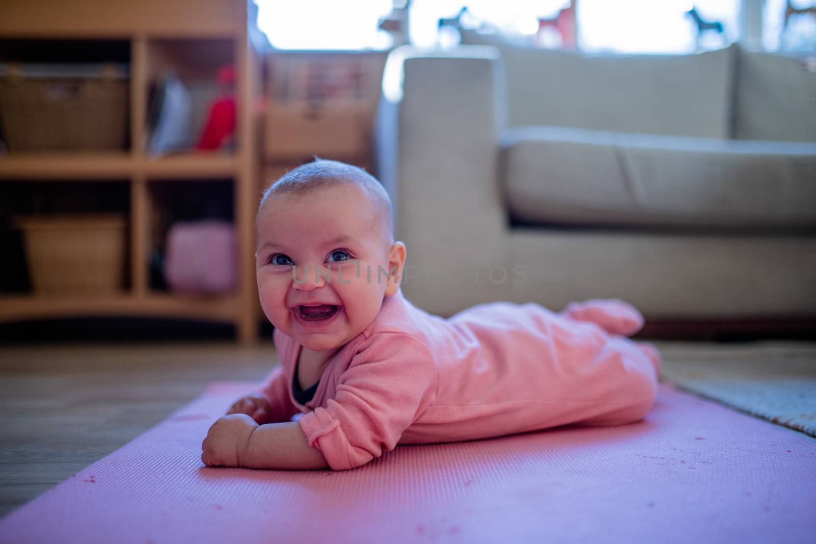 Picture of a smiling baby in a pink sleepsuit lying face down on a pink rug with a couch and a window as background