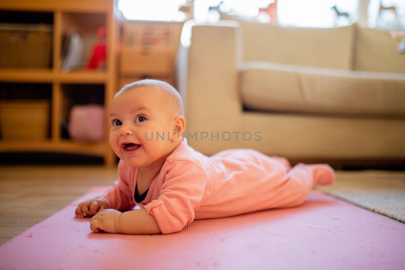 Picture of a smiling baby in a pink sleepsuit lying face down on a pink rug with a couch and a window as background