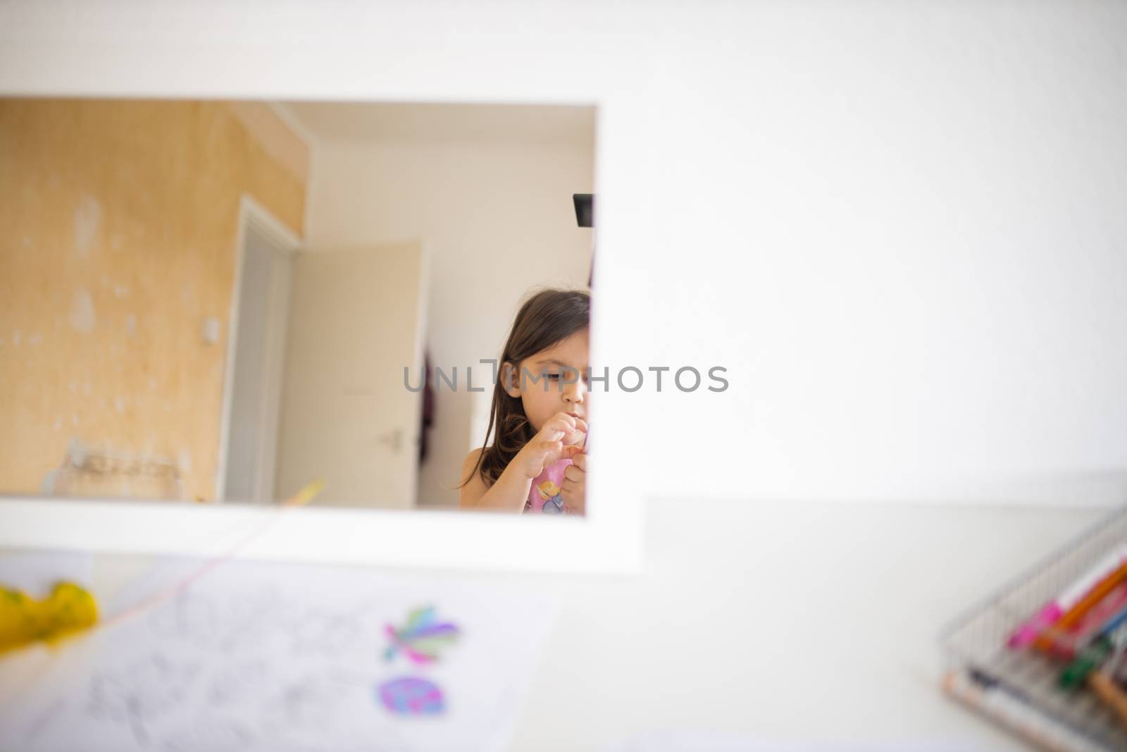 Little brunette girl and a white open door reflected on a mirror across a white table with a drawing of colorful leaves