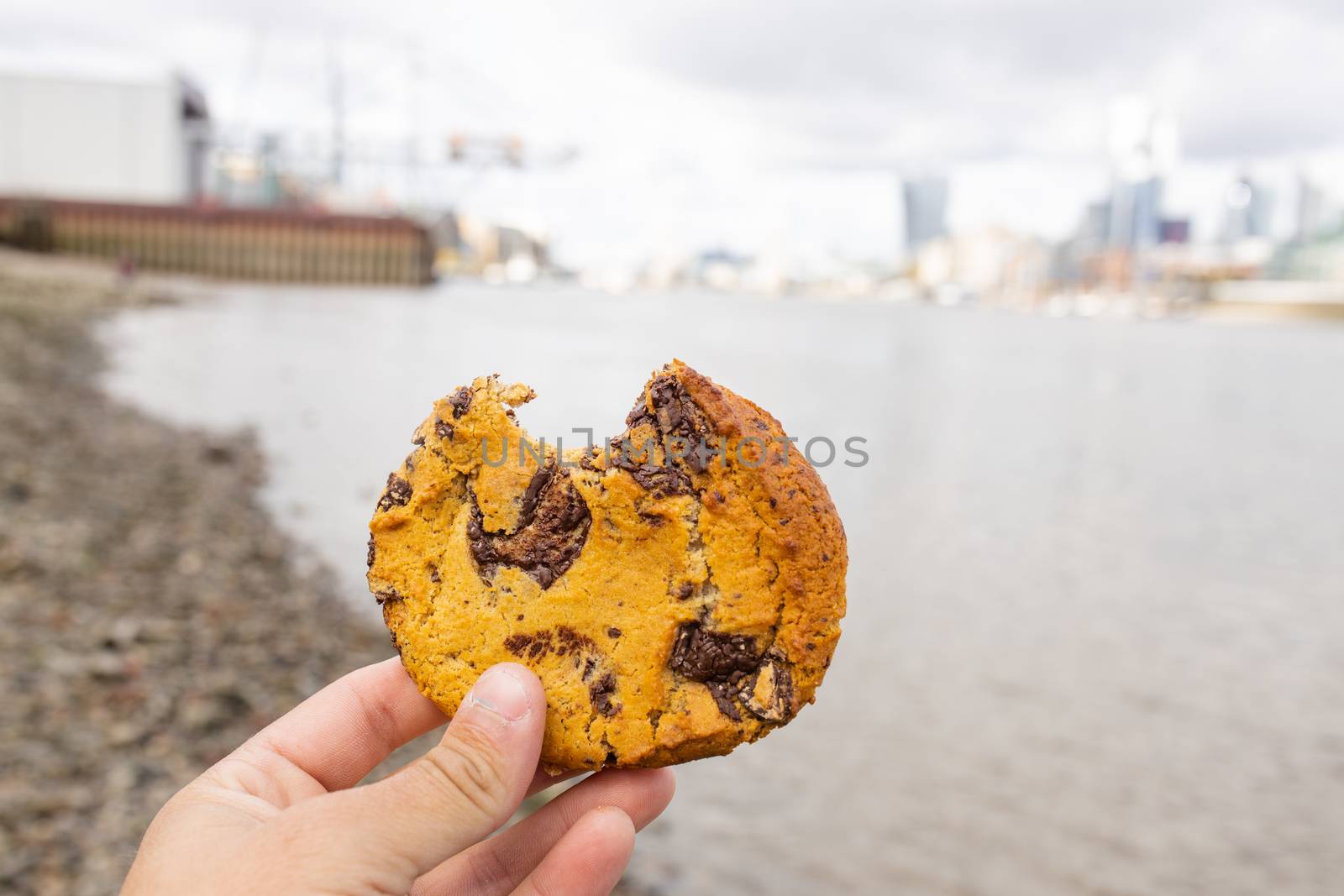 Landscape View of a Hand Holding a Bitten Chocolate Chips Cookie with a Blurry River Thames and CIty as Background