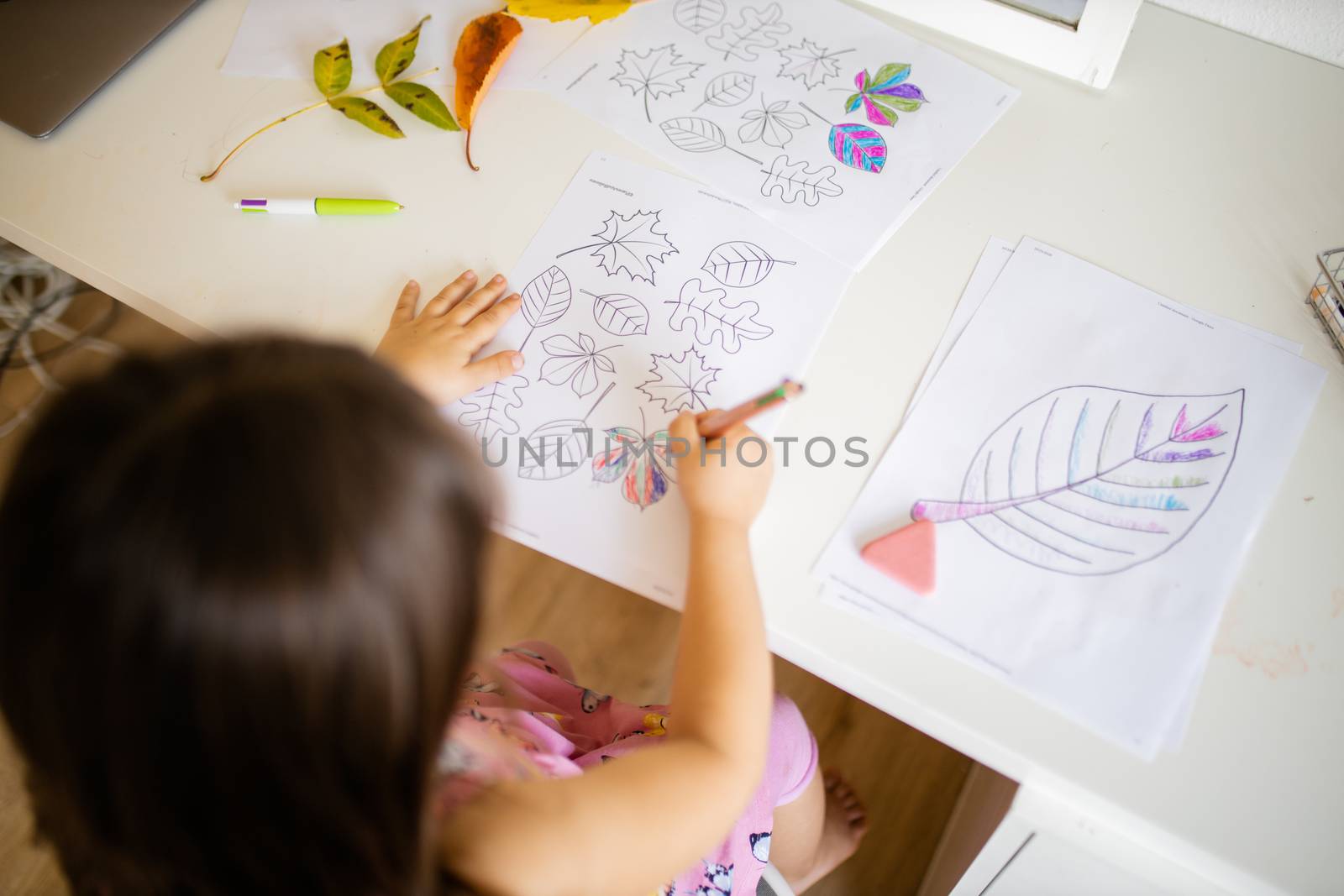 Over the head of a little brunette girl in a pink dress sitting in front of a mirror and coloring leaves