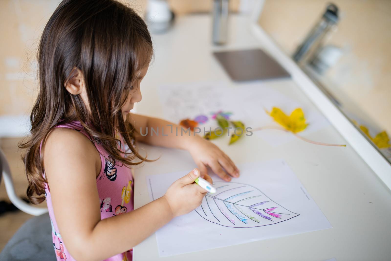 Little brunette girl in a pink dress sitting in front of a mirror and coloring a big leaf image on a paper sheet