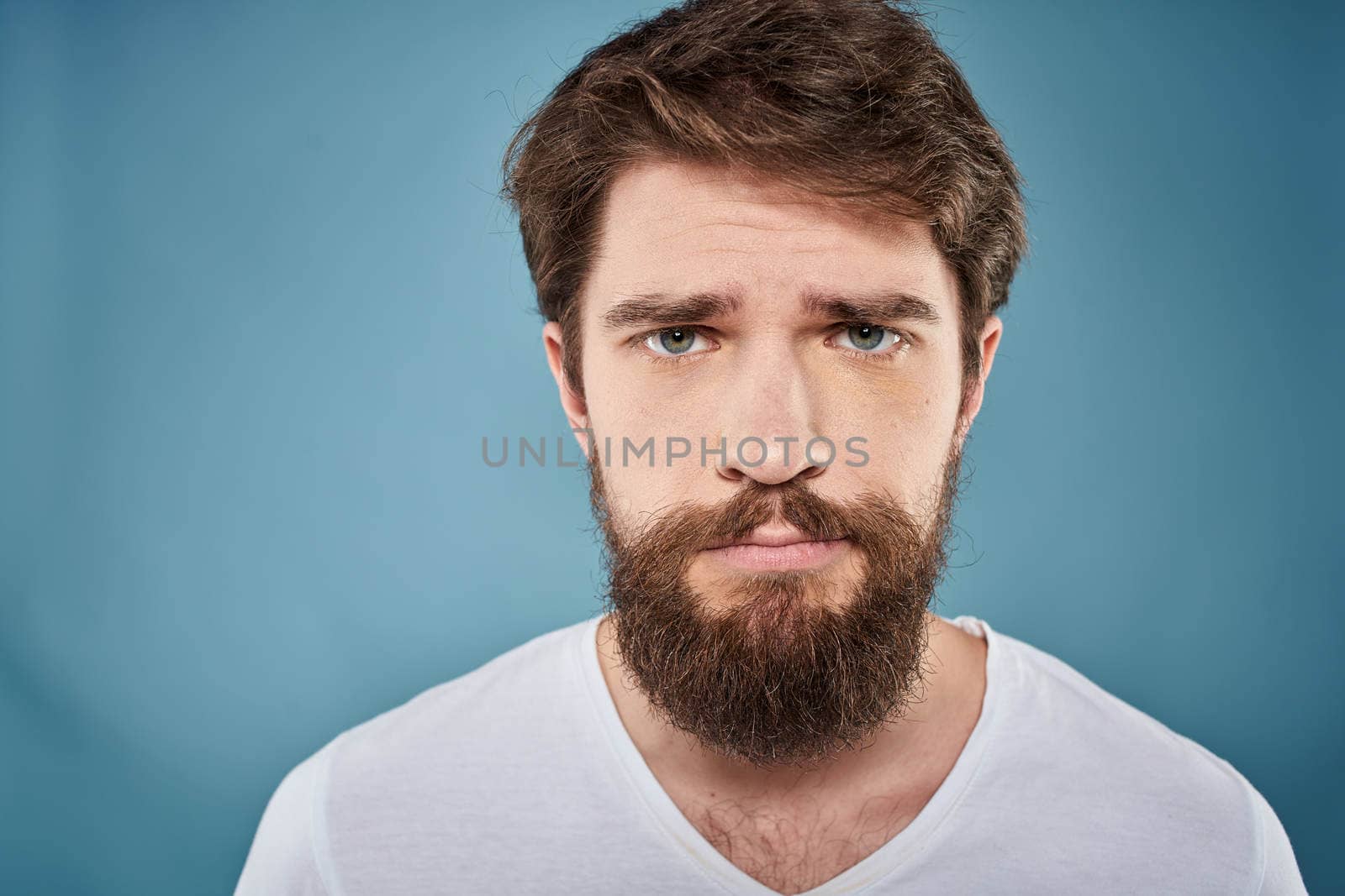 Bearded man displeased facial expression emotions close-up blue background white t-shirt by SHOTPRIME