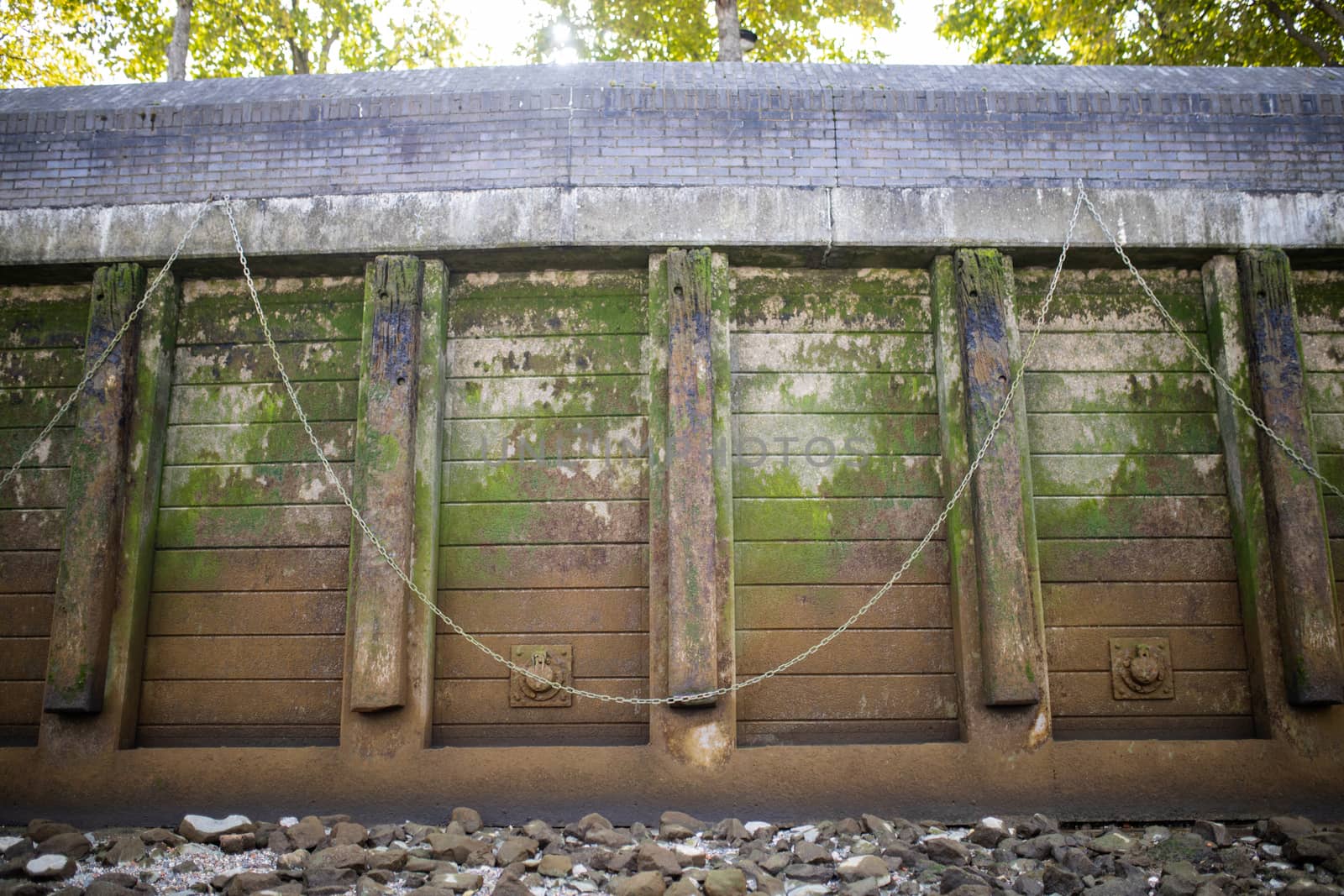 Landscape View of a brown Concrete Wall Partially Covered by Moss, with chains hanging from it, and a ground covered by gray rocks at the bottom