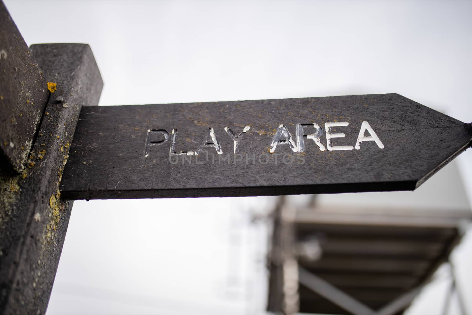 Up close view of a wooden pole with indications pointing out the way to the play area, with a cloudy sky and a blurry structure as background