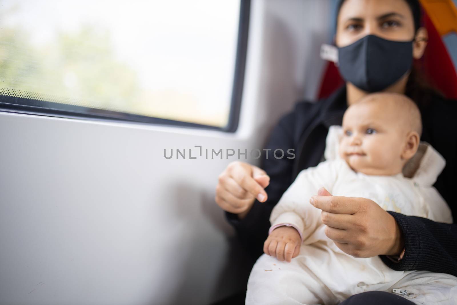 Baby in a white jacket looking through the window of a train while resting on the legs of her mother