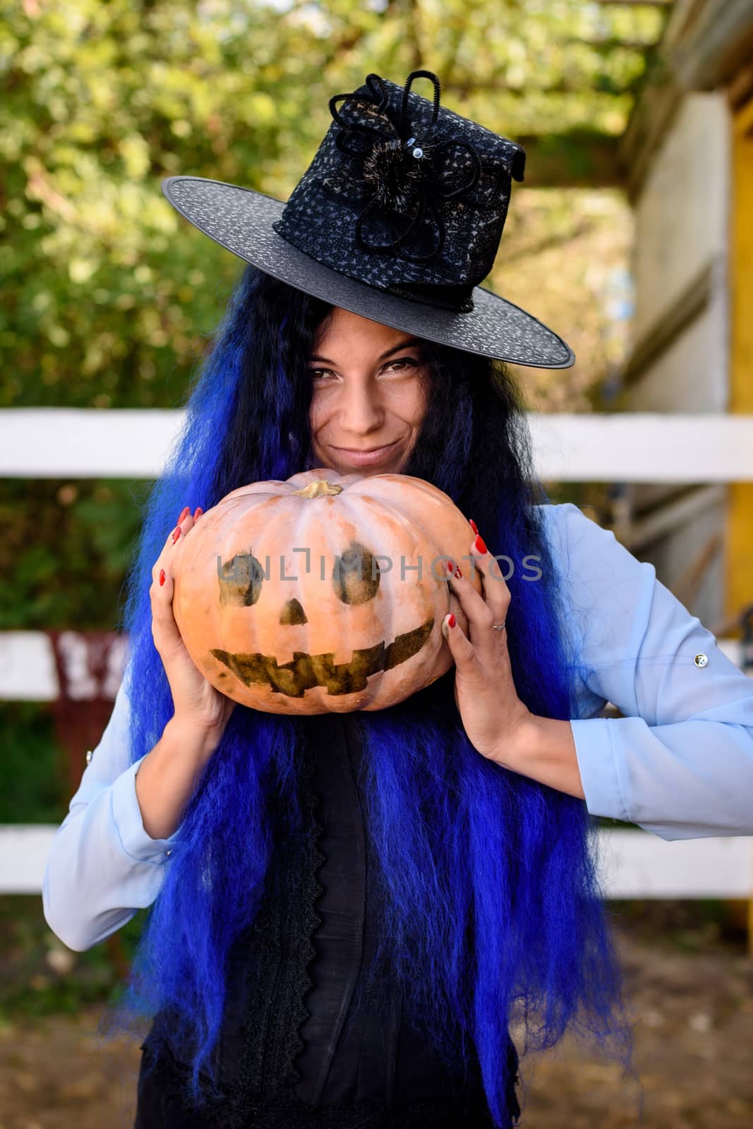 A girl in a witch costume with blue hair is holding a pumpkin with a painted face by Madhourse