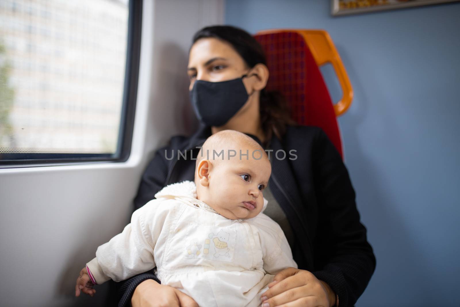 Woman wearing a black face mask sitting next to a window on a train and holding her baby who looks absently