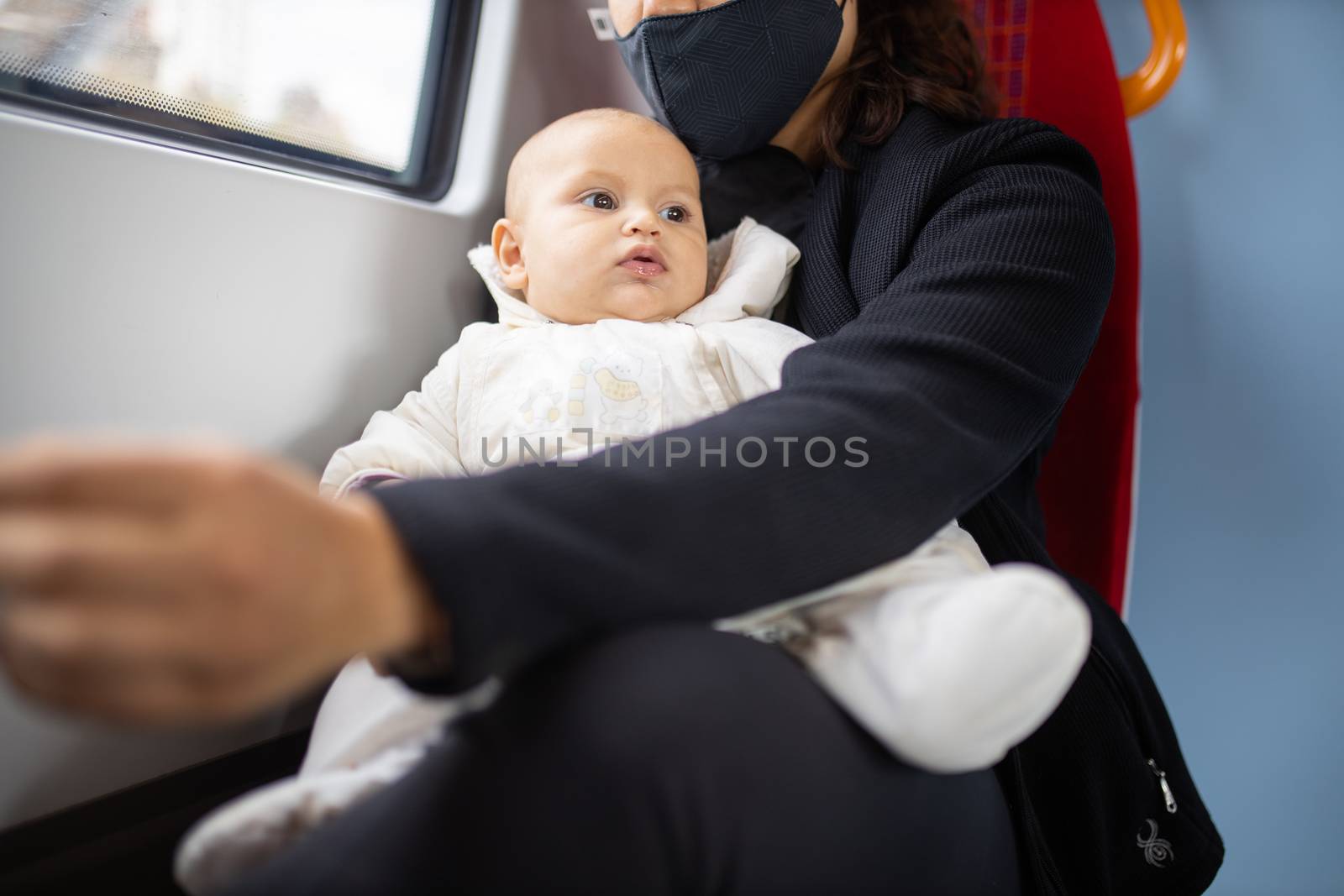 Distracted-looking baby in white clothing resting on the legs of her mother who wears a face mask and sits next to a window