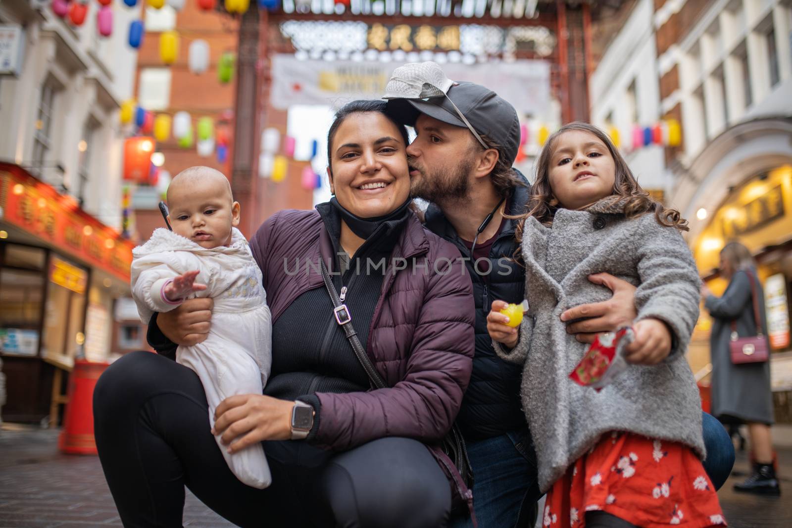 Picture of a smiling woman holding her baby daughter and posing alongside her husband who kisses her while he hugs his daughter, with a blurry Chinese gate from Chinatown as background