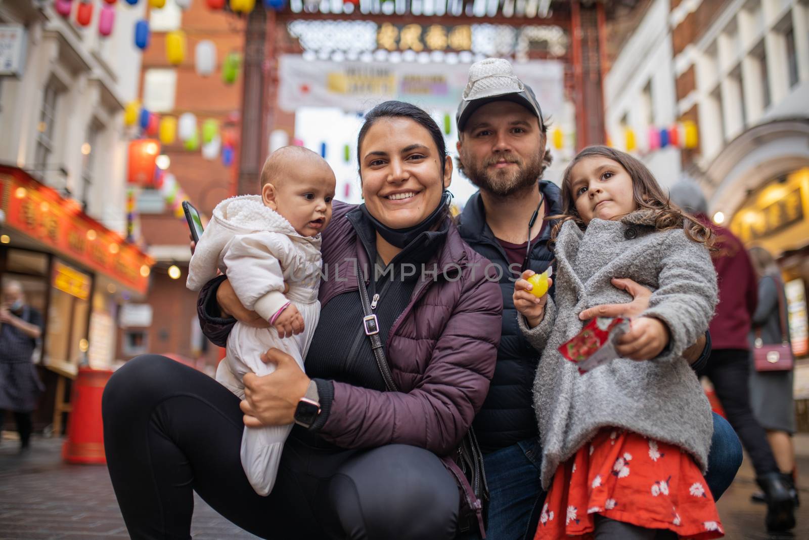 Picture of a smiling woman holding her baby daughter alongside her smiling husband who hugs his daughter, with a blurry Chinese gate from Chinatown as background