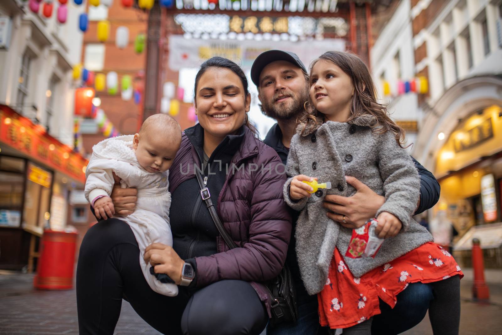 Picture of a smiling woman holding her baby daughter and posing alongside her smiling husband who hugs his daughter, with a blurry Chinese gate from Chinatown as background