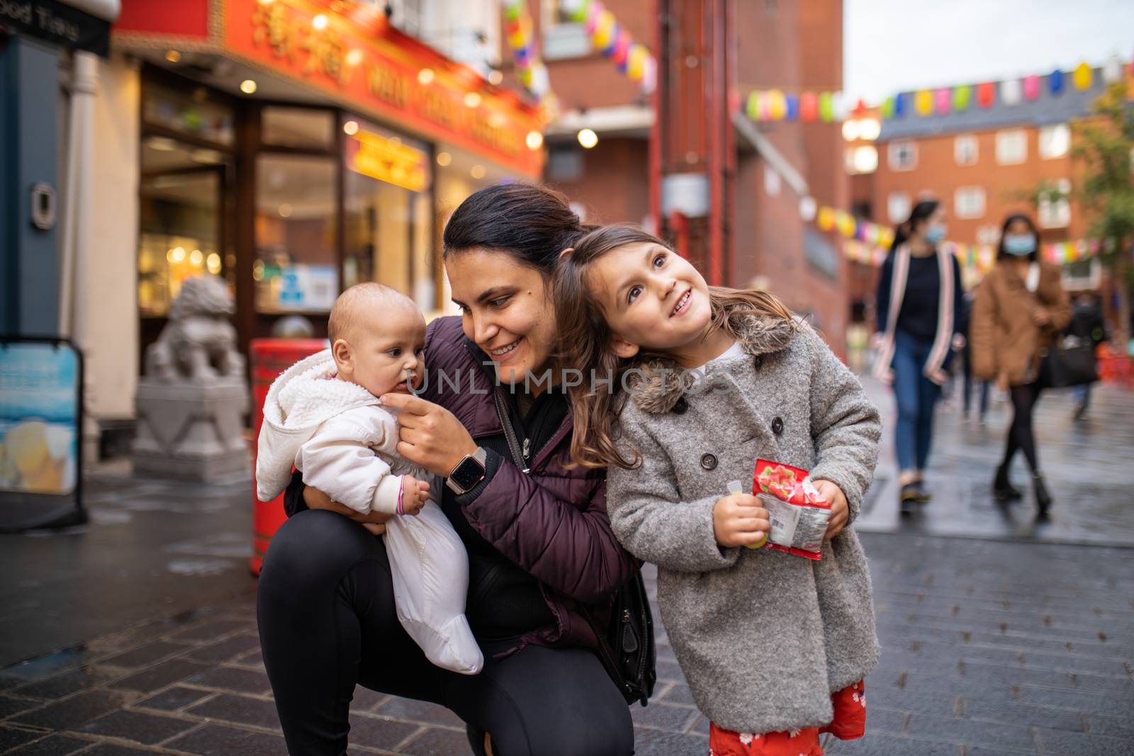 Picture of a smiling little girl resting her head on the shoulder of her mother who is kneeling and holding her baby daughter, with a blurry alley from Chinatown and people walking as the background