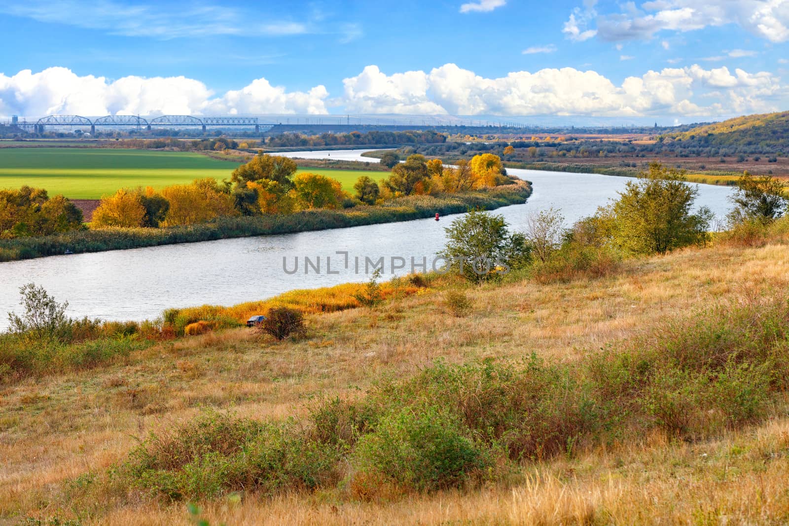 A beautiful autumn landscape with a smooth bend of the river among the plain against the background of a railway bridge on the horizon and a blue sky with white clouds.