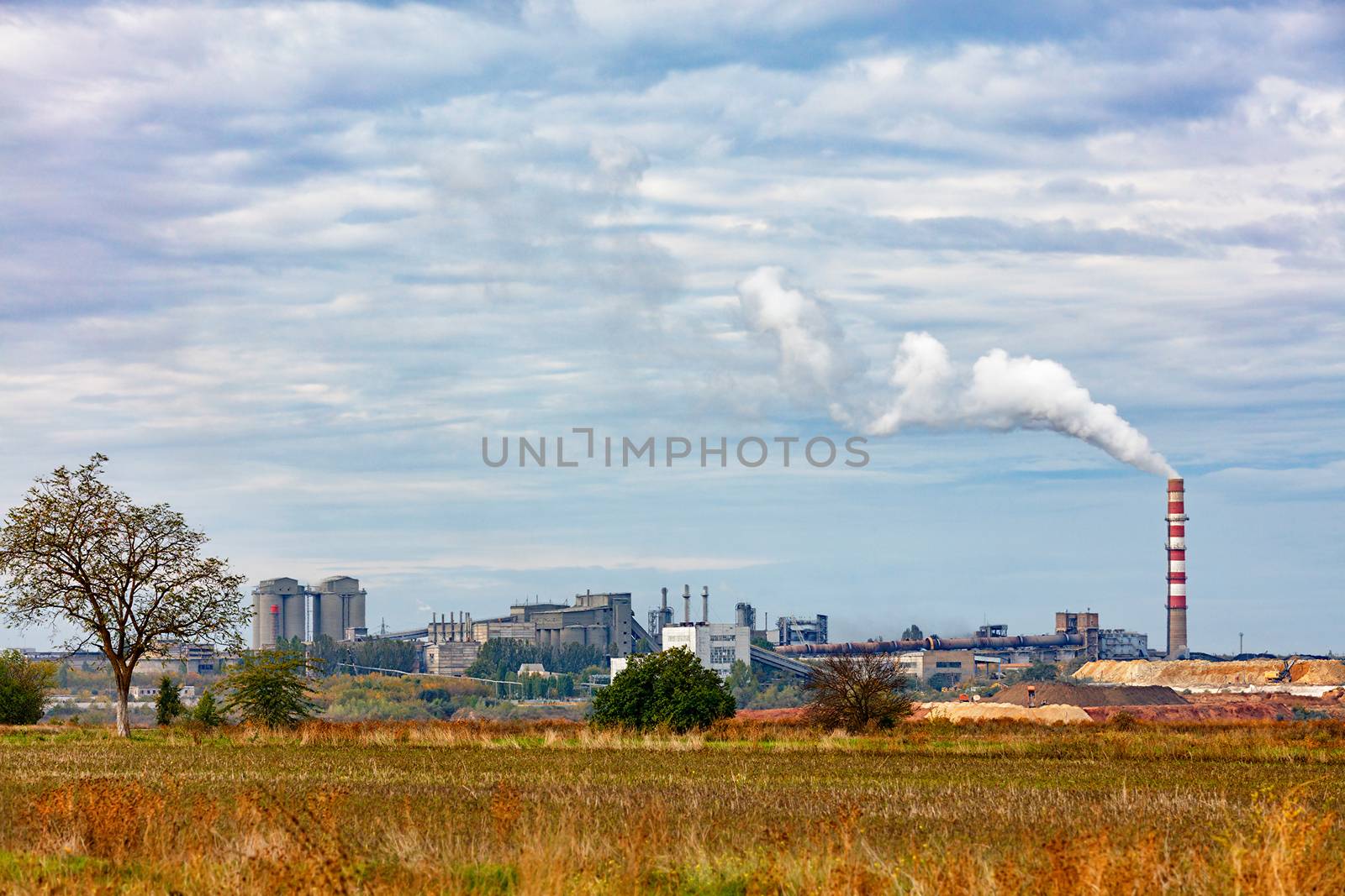 The production complex of a cement plant on the horizon behind an agricultural field. by Sergii