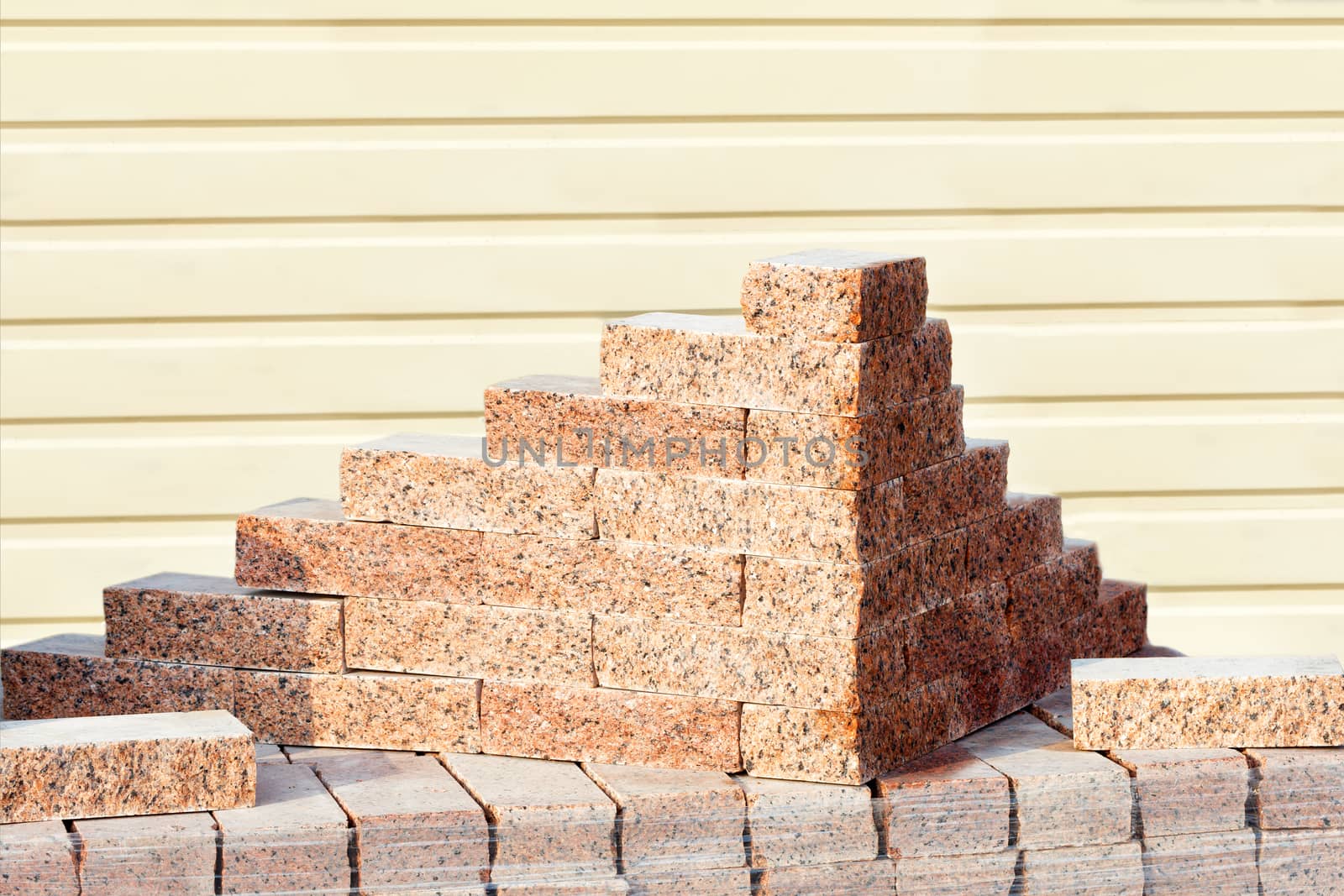 Neat carved red granite blocks stacked outdoors on a bright sunny day against a striped yellow siding wall background, copy space.