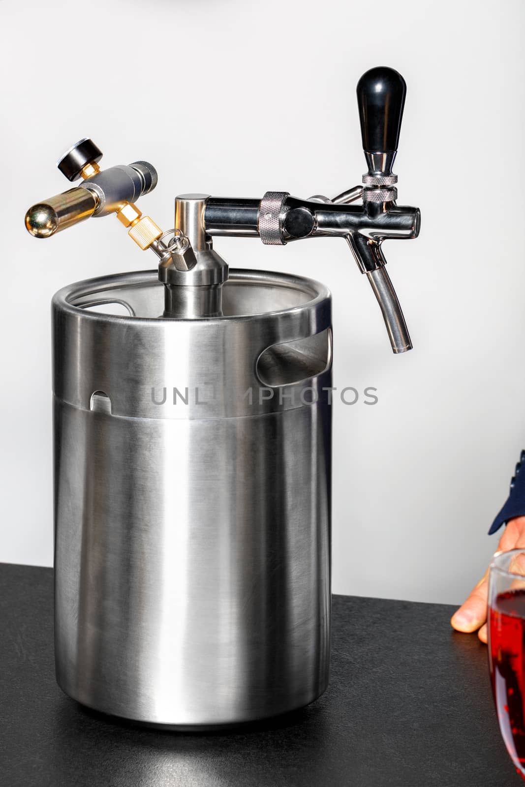 A new clean stainless steel barrel and a tap on it, with a glass of red wine and part of a man's hand visible in the foreground in the corner.