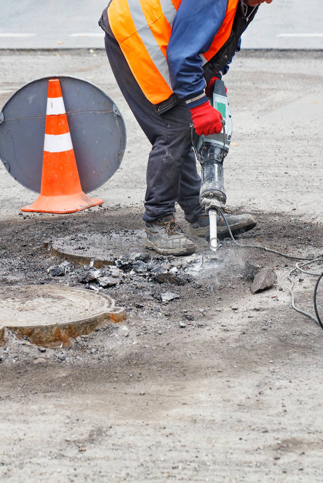 A road worker, dressed in reflective clothing, repairs a section of road near sewers with an electric jackhammer, vertical image, copy space.