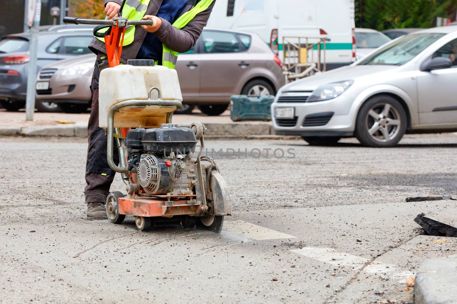 A worker cuts the old asphalt on the road with a gasoline cutter against the background of a city street and passing cars in blur. by Sergii