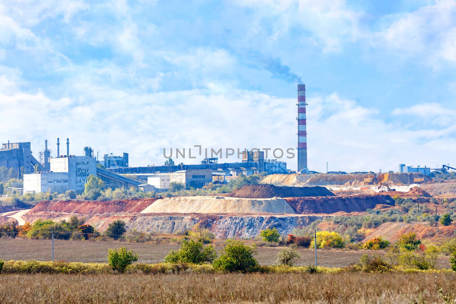 Sand and clay quarry, cement production complex on the horizon with a tall chimney against a blue cloudy sky, copy space.