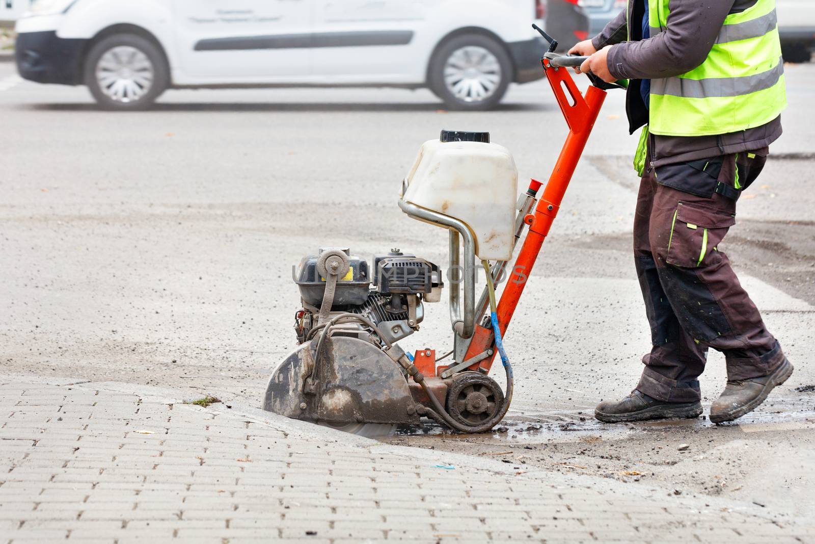 A road worker in a light green reflective vest cuts old asphalt with a petrol cutter on the road in front of a city street in a blurred form. Copy space.