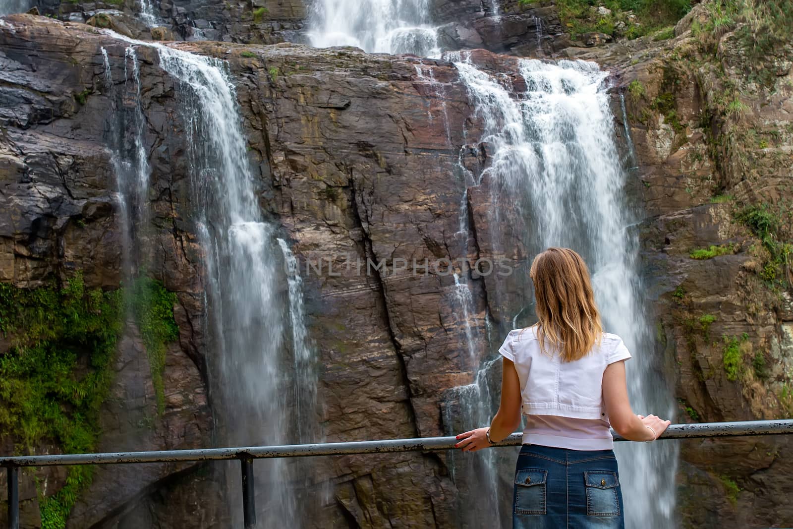 A girl stands in front of a waterfall and admires the beautiful view of falling water. Blond woman on the observation deck and waterfall. Viewpoint