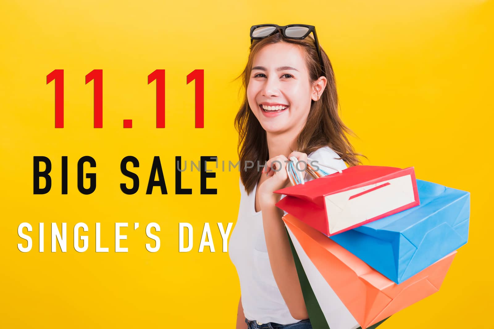 Asian portrait happy beautiful young woman smiling stand with sunglasses excited holding shopping bags multi color with 11.11 big sale and single's day text on space isolated yellow background