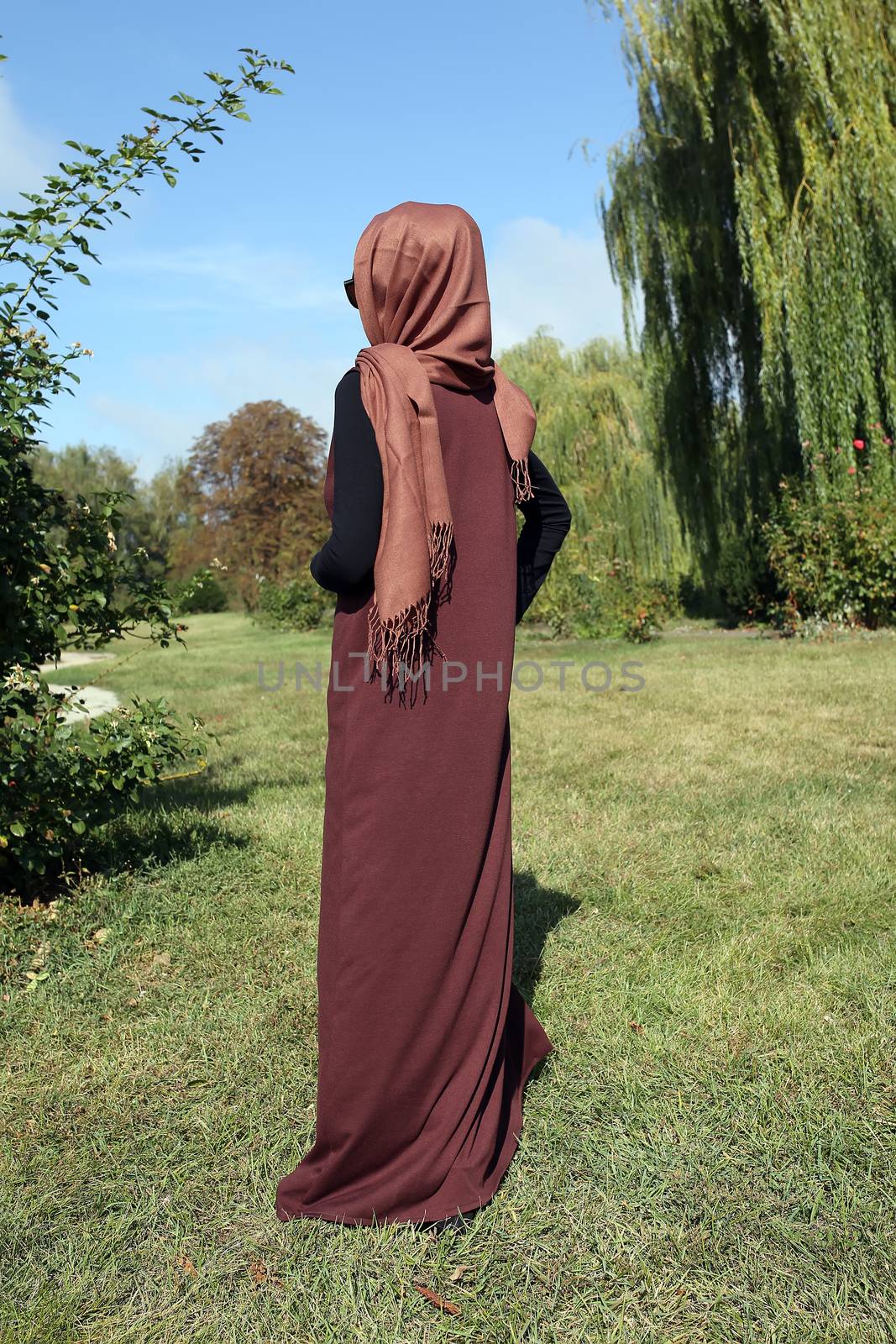 Woman in a hijab. Girl in traditional muslim brown clothes on a background of green grass. Stands with your back.