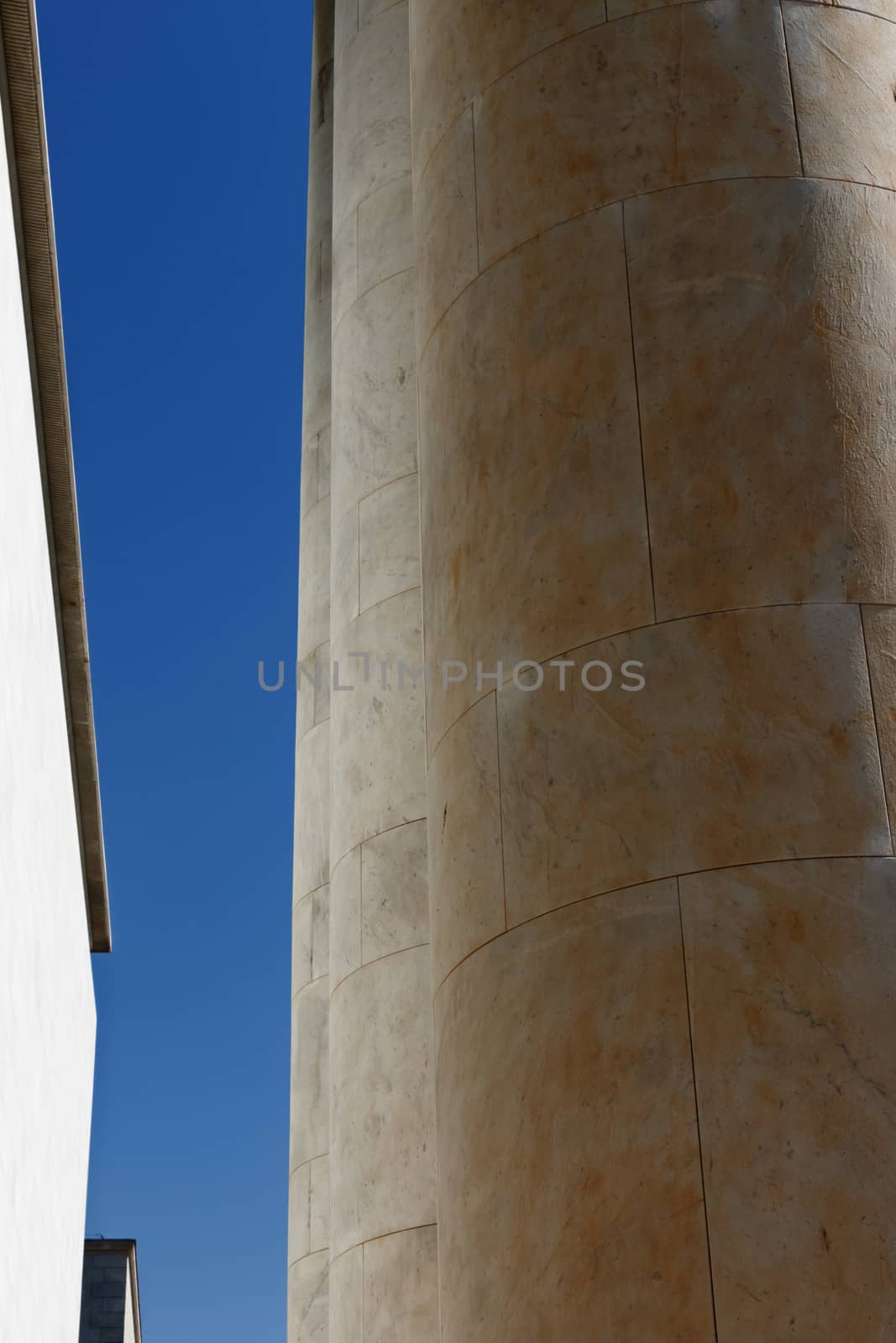 A patch of blue sky between a row of round columns in backlight and a white wall in full sun , vertical composition , wide angle lens from the bottom to the top 