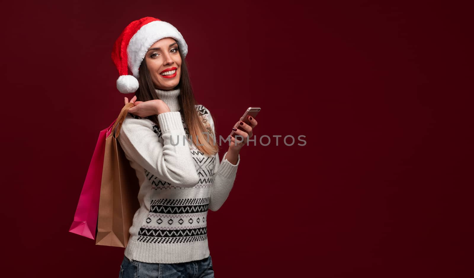 Portrait beautifiul caucasian woman Santa hat on red studio background. Christmas New Year holiday concept. Cute girl teeth smiling positive emotions with shopping bag and smartphone Online shopping