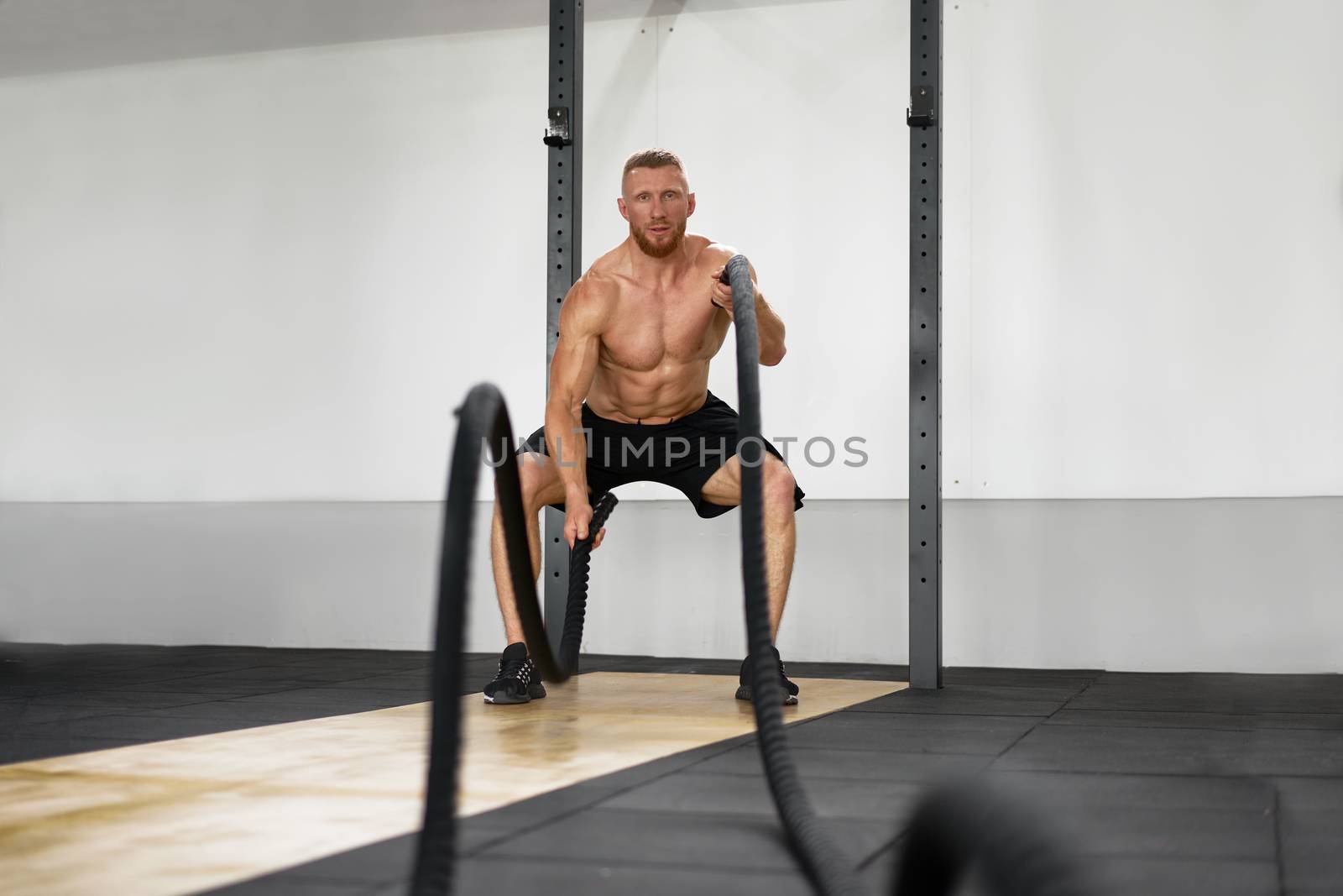 Gym battle rope man stamina training Athlete guy fitness exercising endurance indoor workout. Handsome caucasian sportive guy doing exercise functional training fitness. Muscular men naked torso