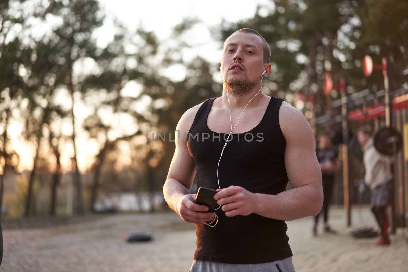Young adult caucasian athlete listening phone music white headphones after workout. Handsome sportsman resting after cross training exercises sunset background. Healthy lifestyle concept.
