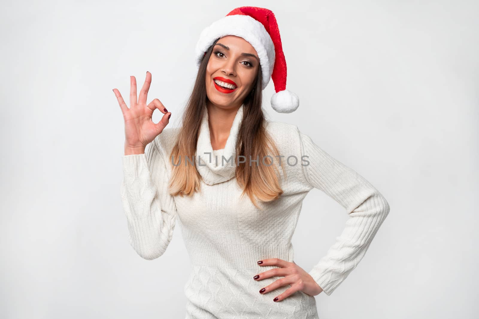 Close up portrait beautifiul caucasian woman in red Santa hat on white studio background. Christmas New Year holiday concept. Cute girl teeth smiling positive Showing ok sign gesture
