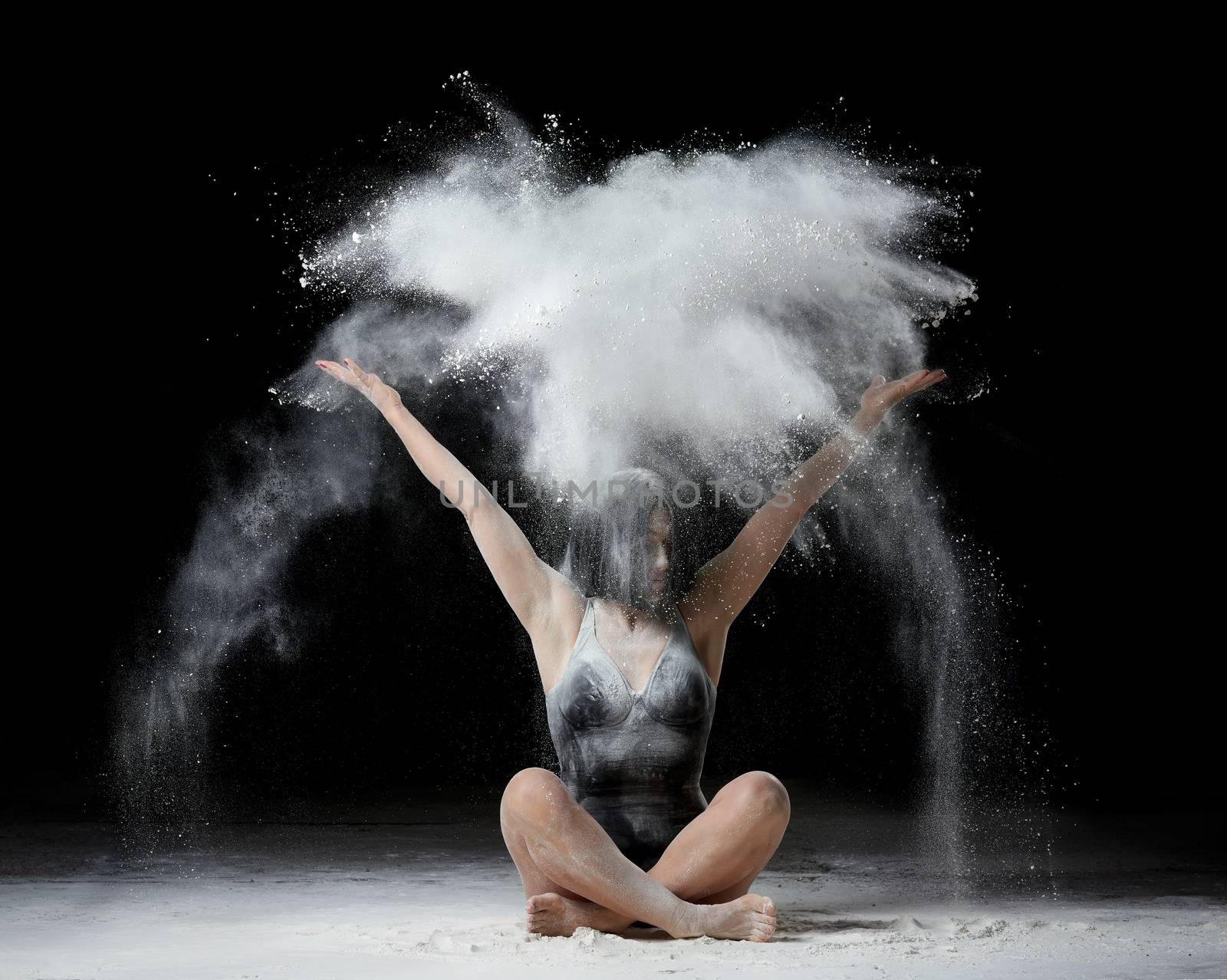 young woman with an athletic body sits on the floor and throws white flour up, dust flies in different directions, athlete is dressed in a black bodysuit