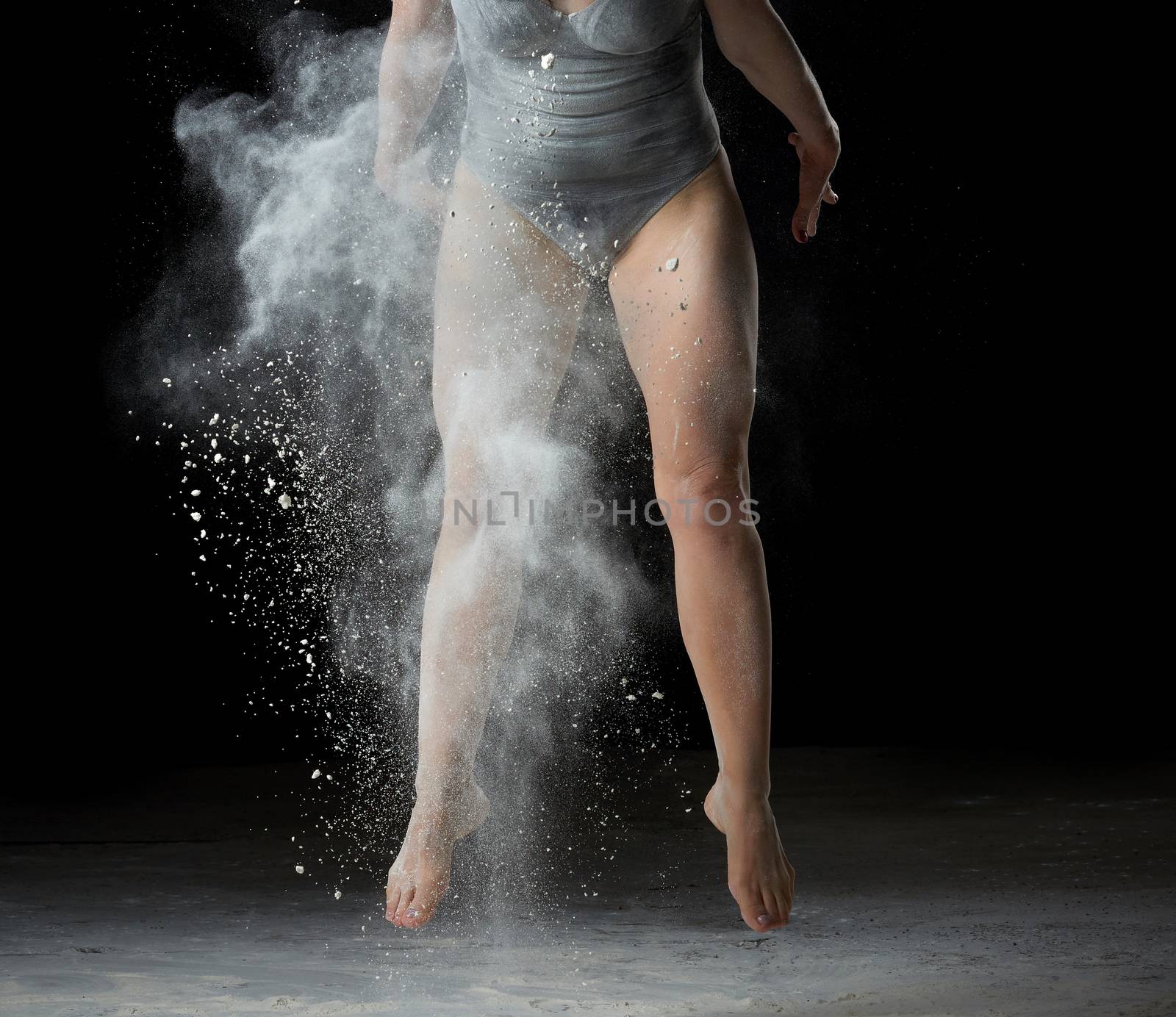 muscular legs of a dancing athlete in a jump, white flour dust cloud, black background, woman dressed in black bodysuit