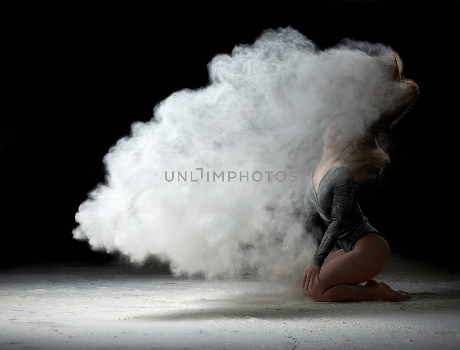 beautiful girl in a black bodysuit with long hair sits and raises her head up, white flour flies in different directions on a black background, explosion