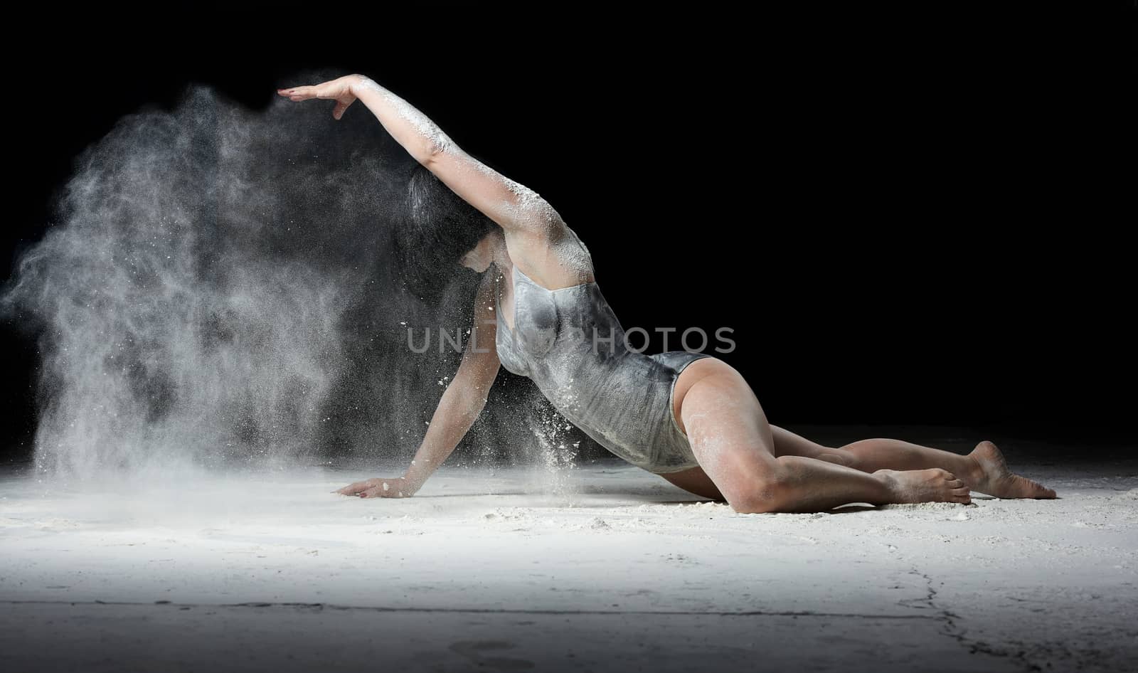 young beautiful woman with black hair is dressed in a sports black bodysuit and lies on the floor and throws white flour over her, black background