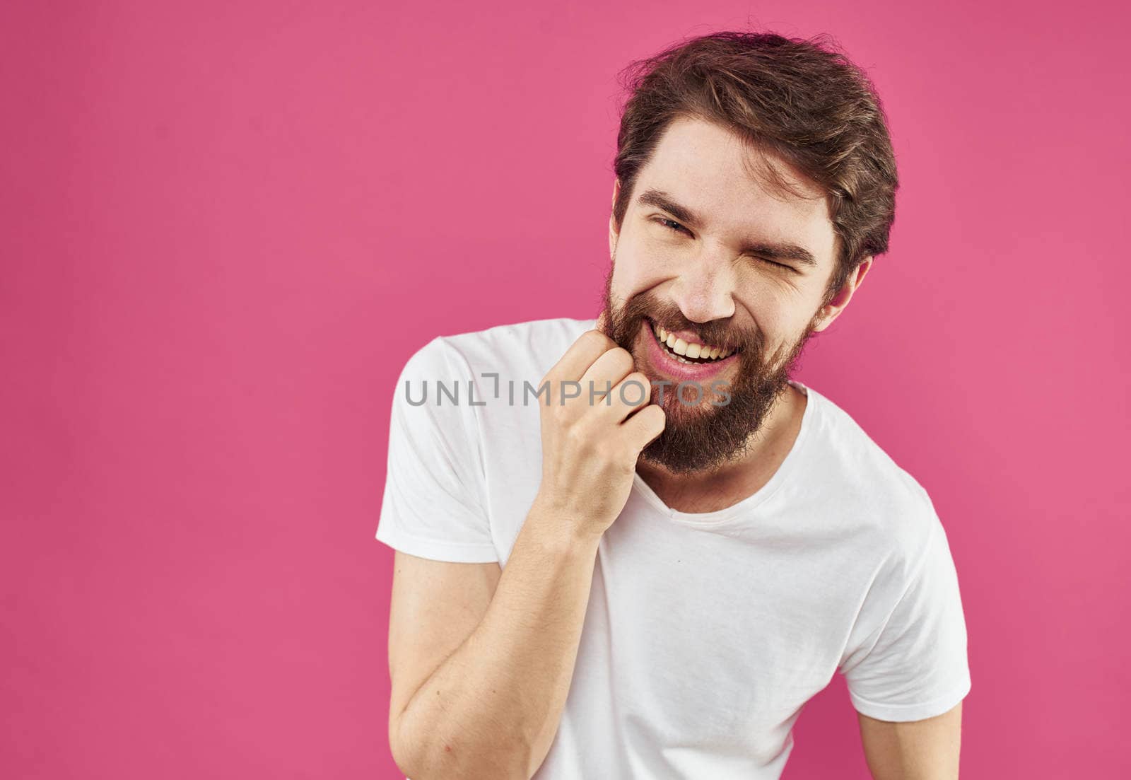 A cheerful guy with a beard laughs on a pink background and gestures with his hands. High quality photo