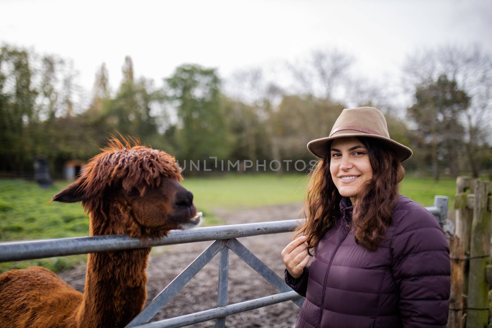 Smiling brunette woman wearing a hat and standing next to a brown alpaca while looking at the camera, and with a blurry forest and cloudy sky as background