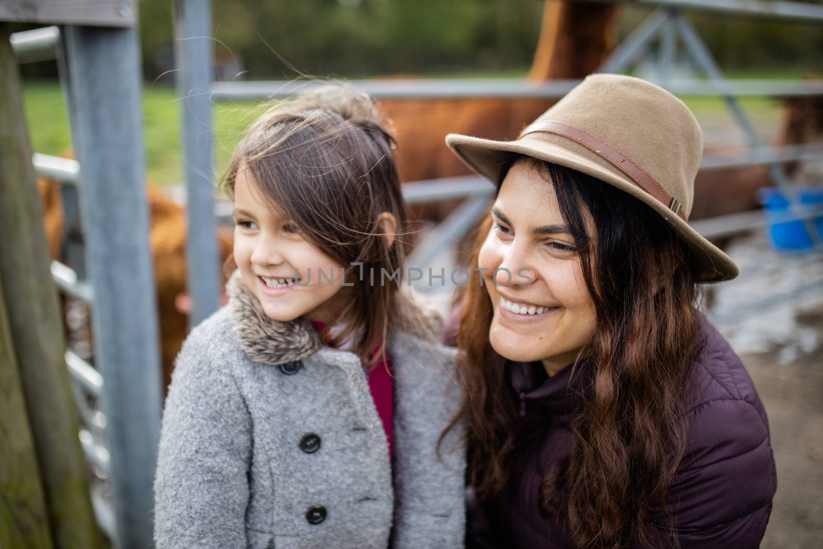 Happy brunette woman and her little daughter smiling and looking at the distance, while brown alpacas eat behind them
