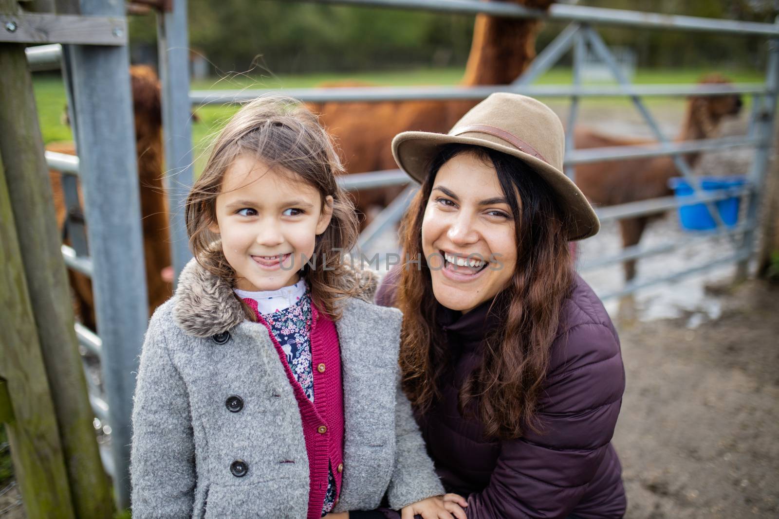 Mother and daughter smiling at the camera with brown alpacas behind them by Kanelbulle