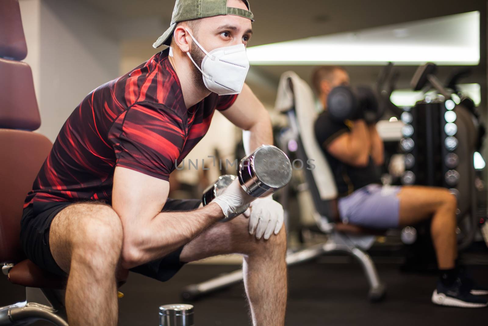 Young men working out wearing face mask & latex rubber gloves,performing bicep curl with dumbbells,COVID-19 pandemic social distancing rules while working out in reopened indoor gym,prevent & protect