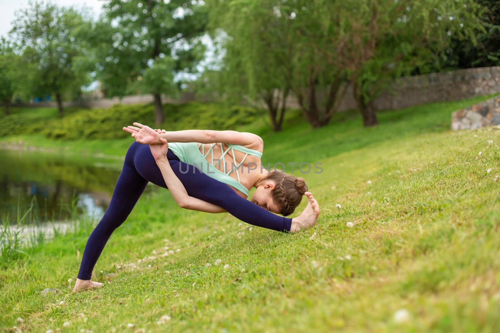 Slender young brunette yogi performs challenging yoga exercises on the green grass in the summer against the backdrop of nature.