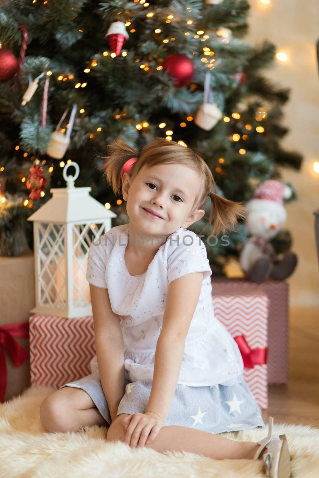 Pretty little child is sitting in front of christmas tree among garlands and presents..