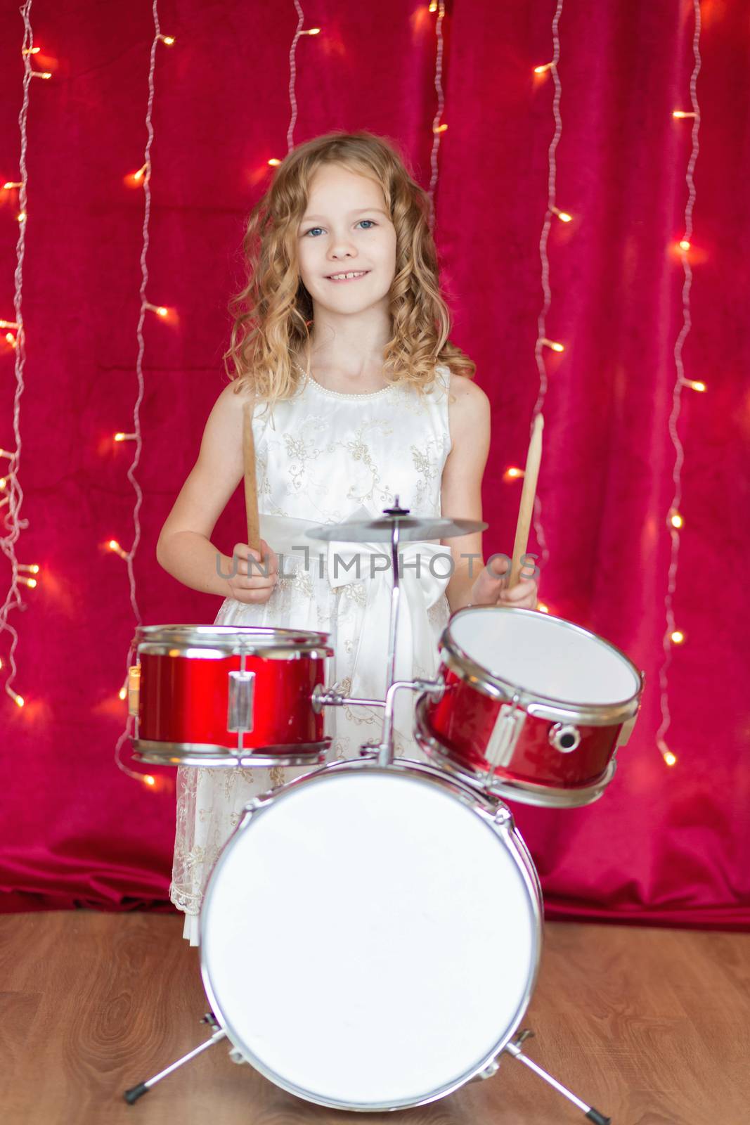 Little smiling girl plays on drums on red background with new year garlands.