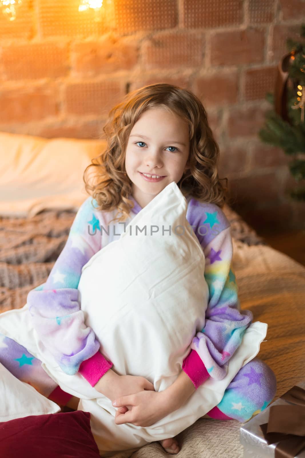 Cute little girl sits on the bed and huggs pillows with background of christmas tree behaind.