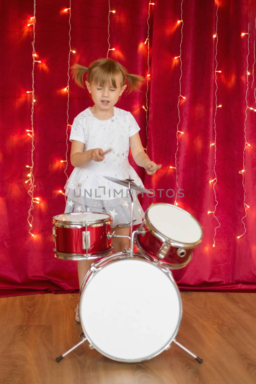 Little smiling girl plays on drums on red background with new year garlands by galinasharapova